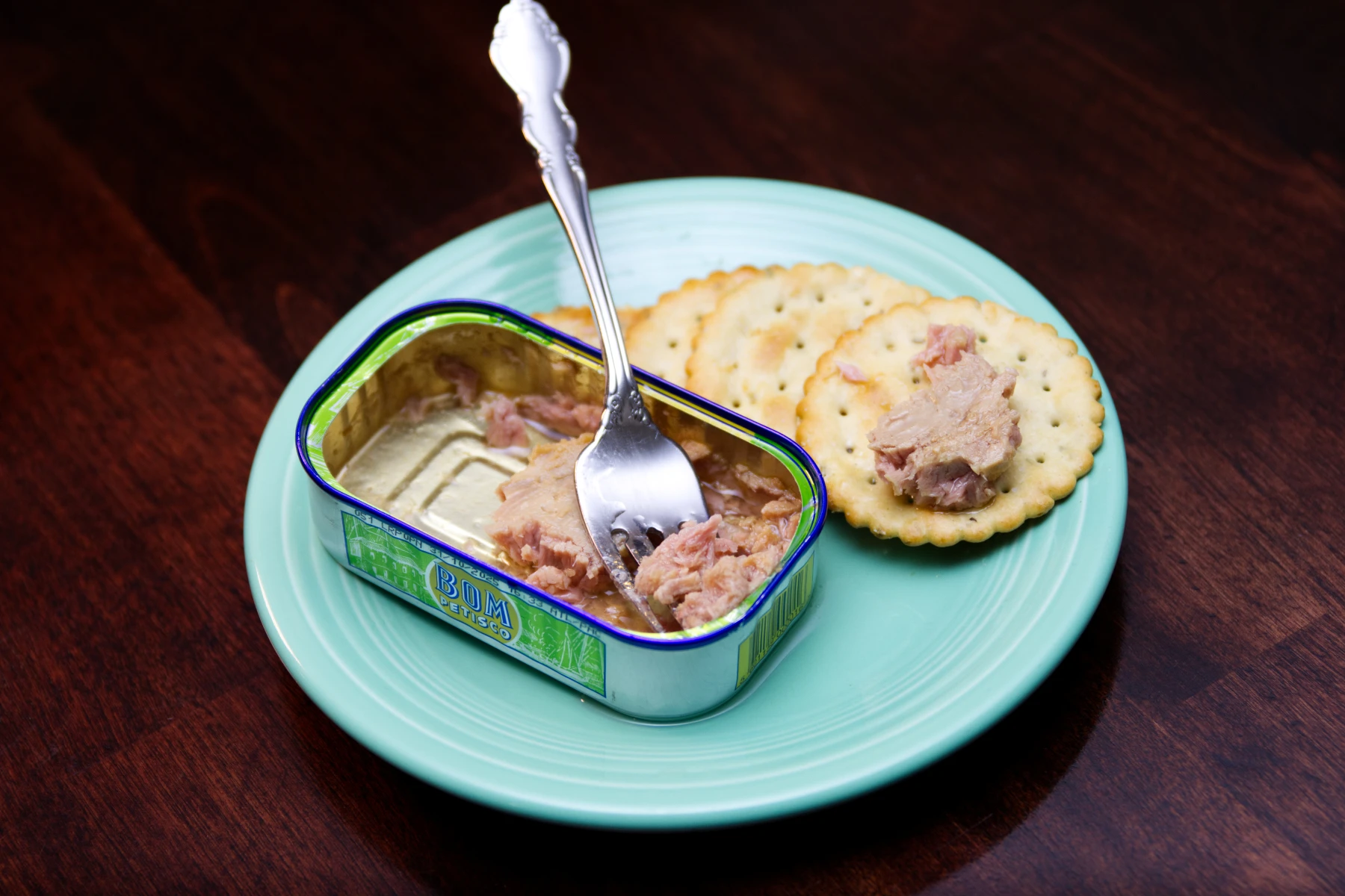 Half empty tin of tuna and a couple of crackers left on a plate. One cracker has a piece of tuna on it.