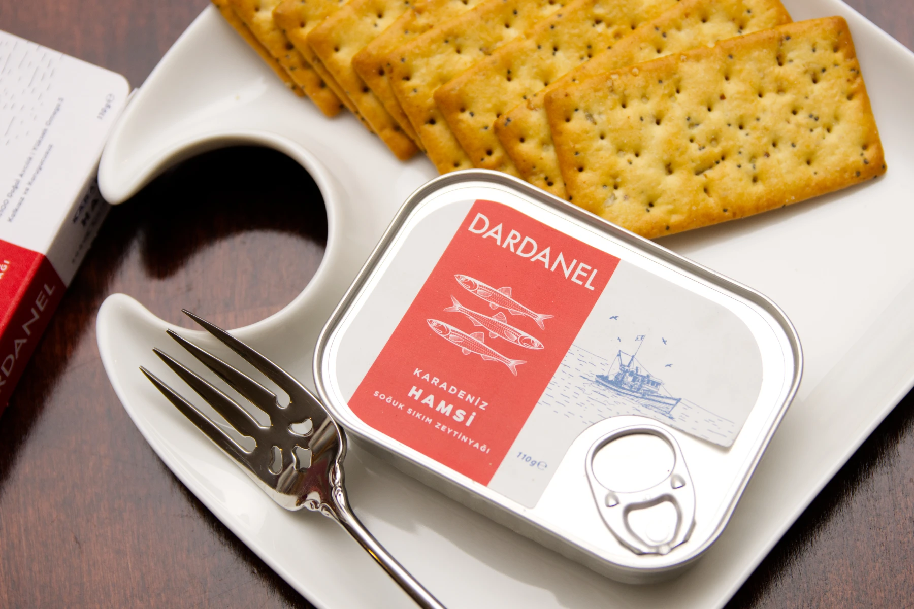 Tin of Dardanel anchovies on a plate with crackers. There is a sticker label adhered to the lid of the tin.