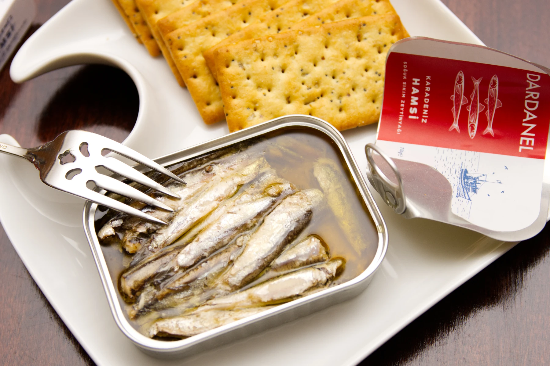 Tin of opened anchovies on a plate with crackers. Fish is stacked in pile in a pool of olive oil.