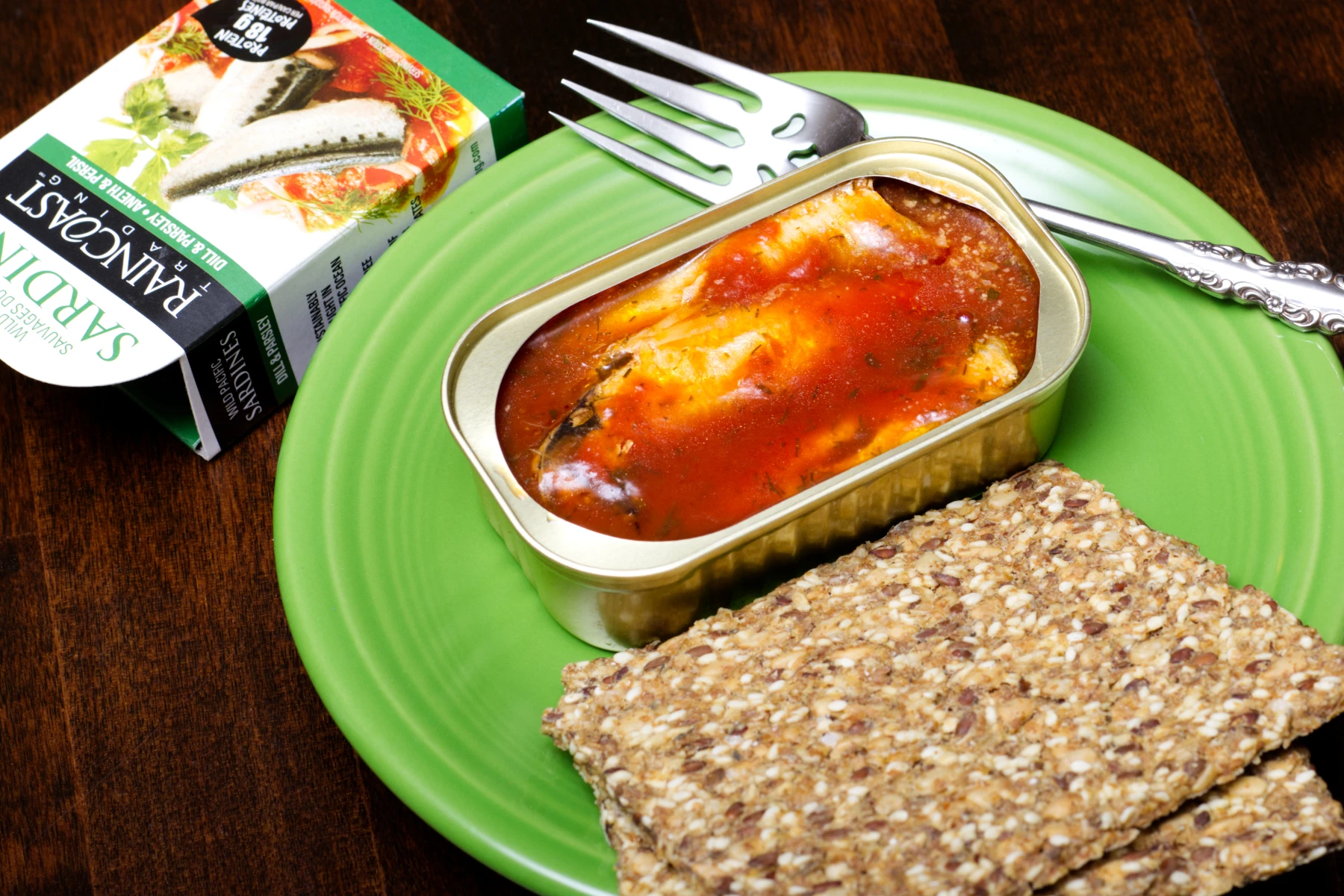 Opened can of sardines, three pieces in an orange red tomato sauce. Two pieces of crispbread on a plate.